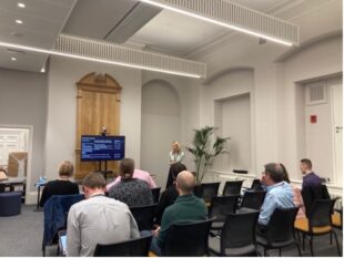 colleagues sat in rows of chairs the OAB event space watching an event. The presenter stood at the front looking at a large screen. Several chairs are empty.