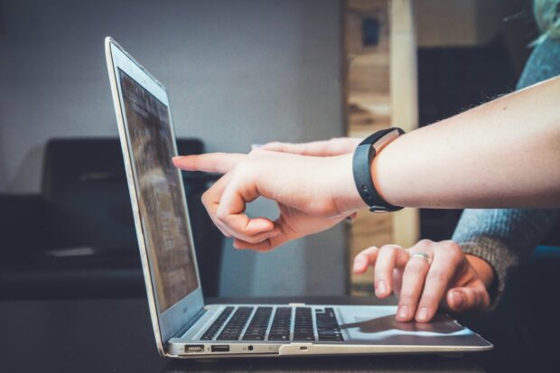 A hand points to a screen on a laptop while another hand scrolls uses the trackpad