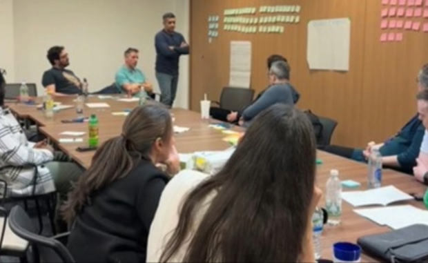 A group of 10 people sat around a table looking at post it stickers on a wall.