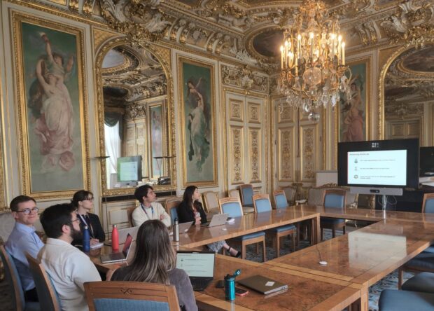A group of 6 people sat around a table in a very ornate room looking at a presentation on a big screen.