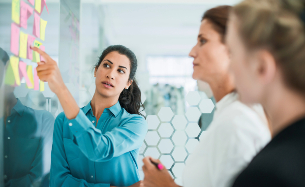 A group of 3 women examine post it notes stuck to a glass wall.