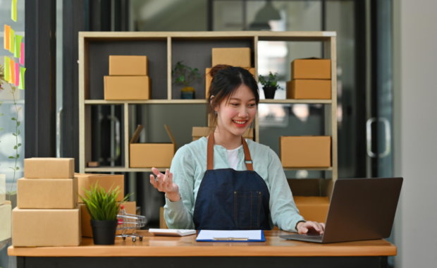 A small business owner is looking down at her laptop and smiling. She is surrounded by small boxes.